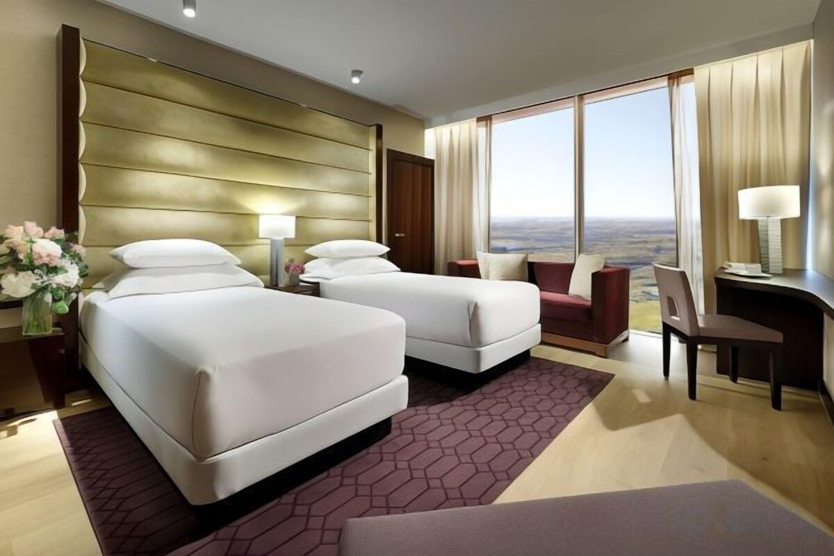 Ein Hotelzimmer mit zwei Betten, einem Schreibtisch, einem Stuhl und einem großen Fenster mit Blick auf die Stadt.