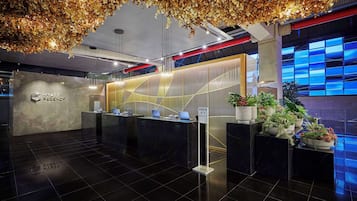Un área de recepción moderna de hotel con mostrador de check-in, pantalla digital y cartel con el nombre del hotel.