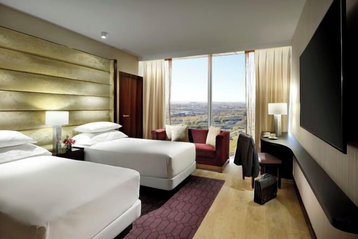 Ein Hotelzimmer mit zwei Betten, einem Schreibtisch, einem Fernseher und einem großen Fenster mit Blick auf die Stadt.