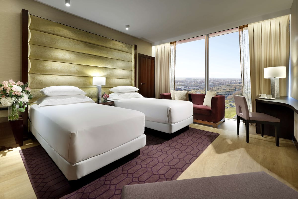 A hotel room with two beds, a desk, a chair, and a large window with a city view.