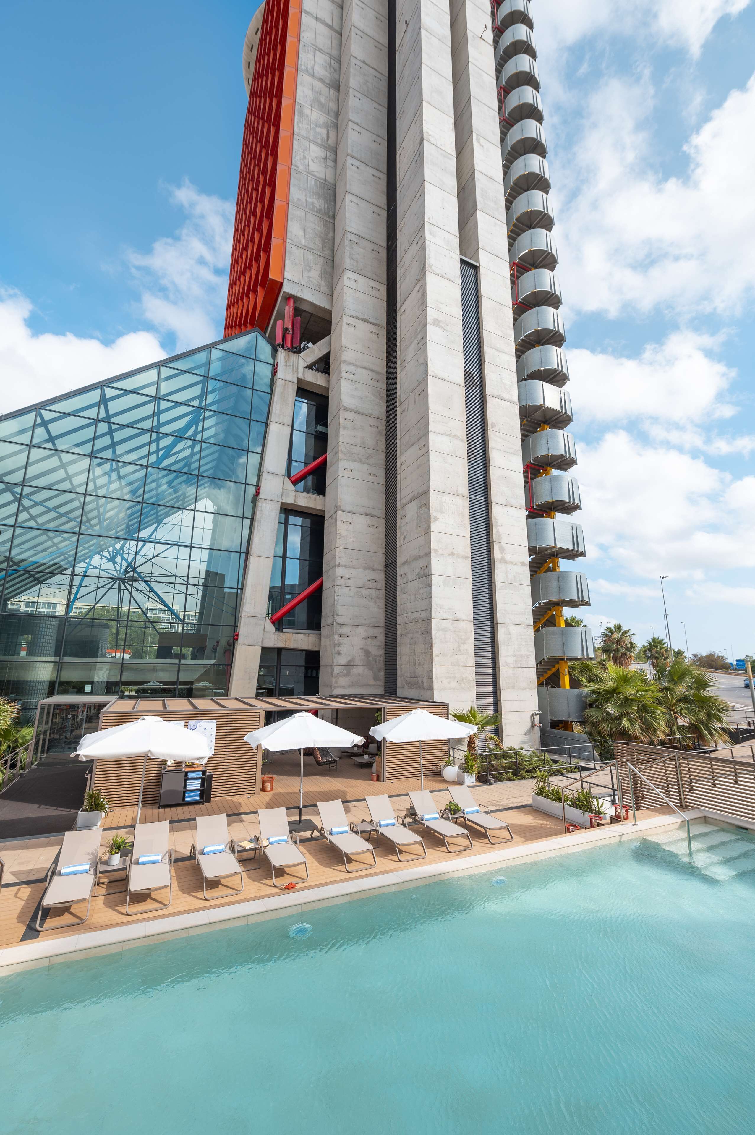 Un edificio moderno con fachada de vidrio y zona de piscina con tumbonas y sombrillas.