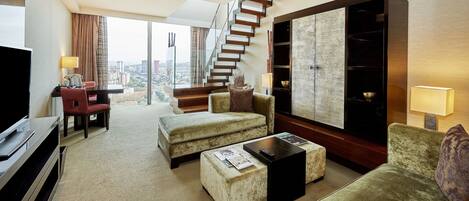 A hotel room with a large window, a sofa, a coffee table, a TV, and a staircase.