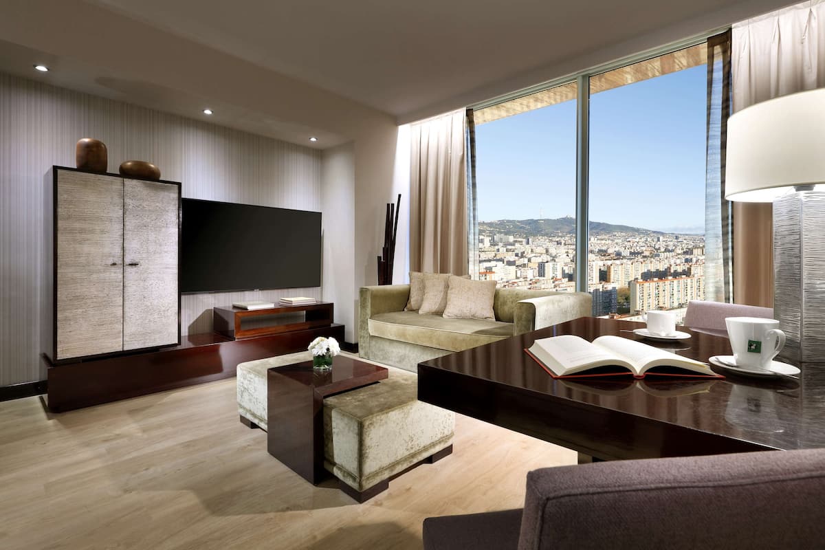 A modern hotel room with a large window offering a city view, a sofa, a coffee table, and a flat-screen TV.