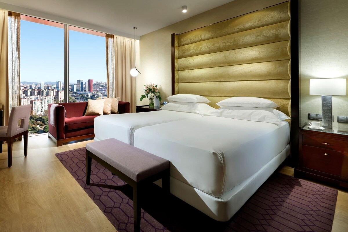Ein Hotelzimmer mit einem großen Bett, einem Schreibtisch, einem Sessel und Blick auf die Stadt.