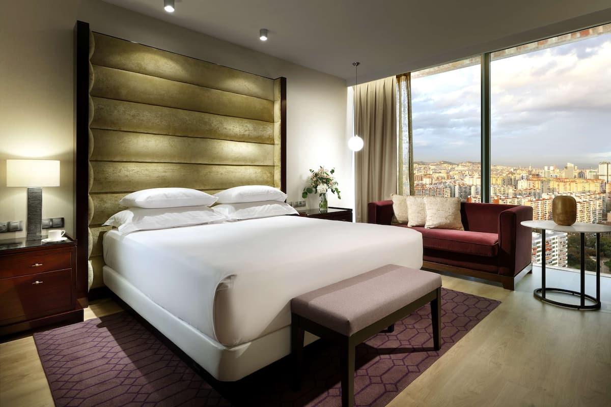 A modern hotel room with a large bed, a bench, a red sofa, a small table, and a city view through large windows.