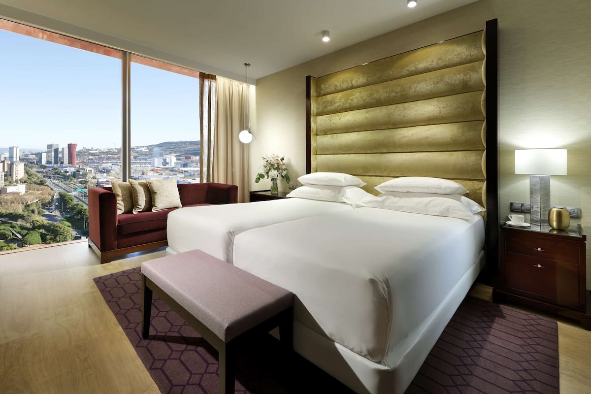 A hotel room with a large bed, a bench, a nightstand, a lamp, and a view of the city.
