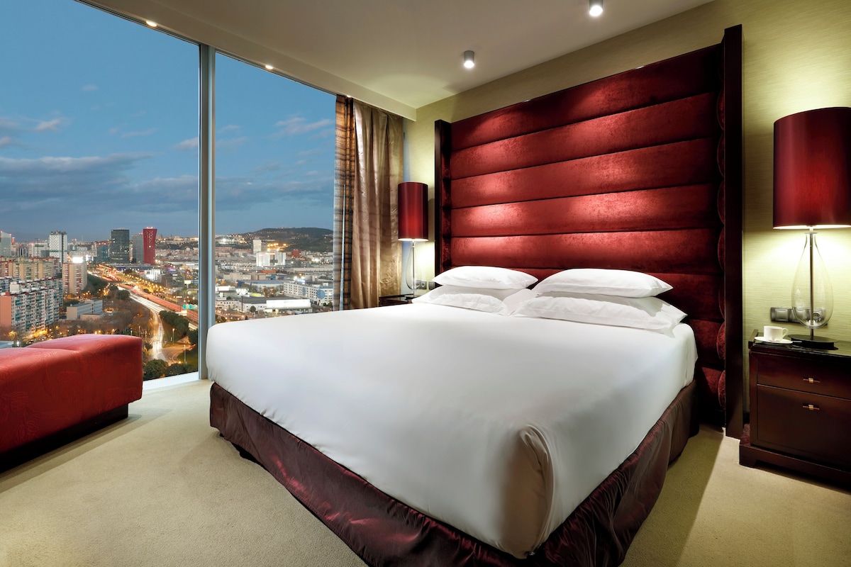 A hotel room with a large bed, a red upholstered headboard, a city view, and a red bench.