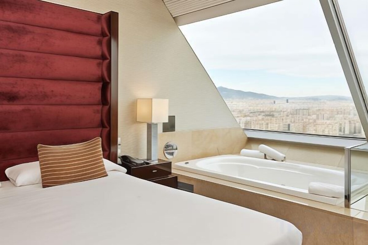 Ein Hotelzimmer mit Bett, Nachttisch, Lampe, Badewanne und Blick auf die Stadt.