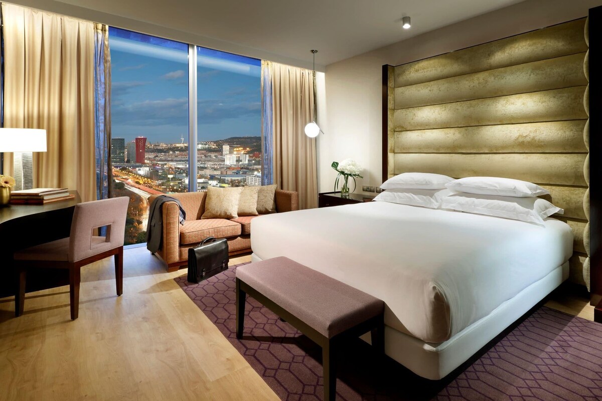 Ein Hotelzimmer mit einem großen Bett, einem Schreibtisch, einem Sessel, einer Bank und Blick auf eine Stadtlandschaft durch große Fenster.