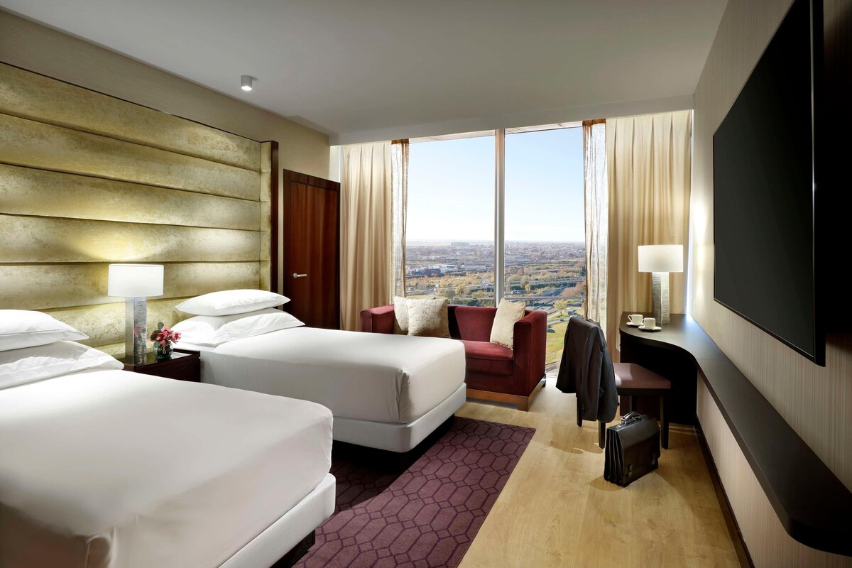 Ein Hotelzimmer mit zwei Betten, einem Schreibtisch, einem Fernseher und einem großen Fenster mit Blick auf die Stadt.