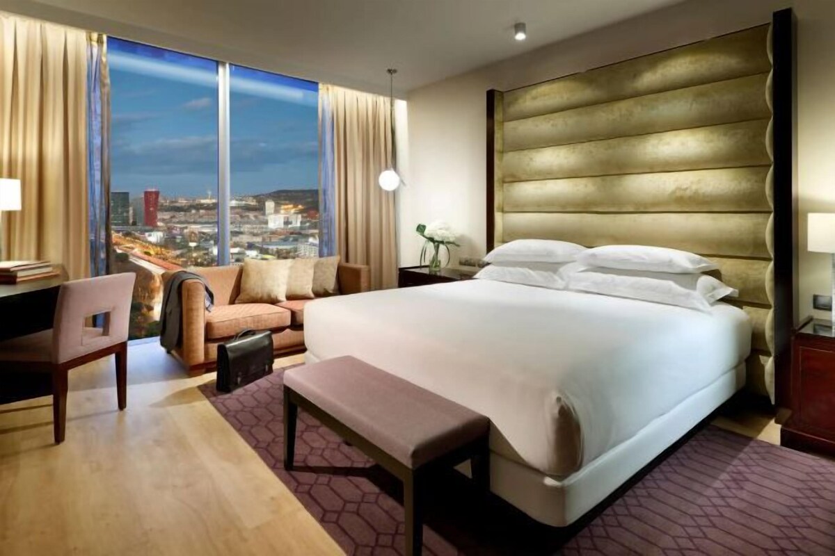A hotel room with a large bed, a desk, a chair, a bench, and a view of a cityscape.