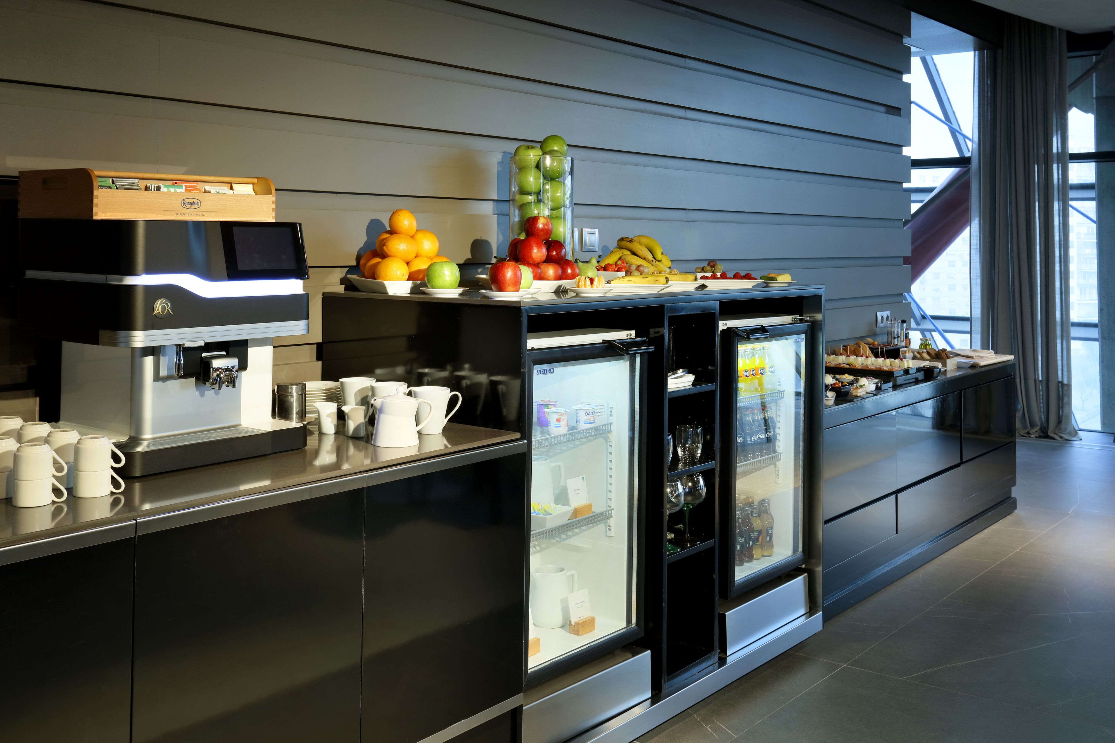 Un área de cocina moderna con una cafetera, una bandeja de fruta y estantes refrigerados.