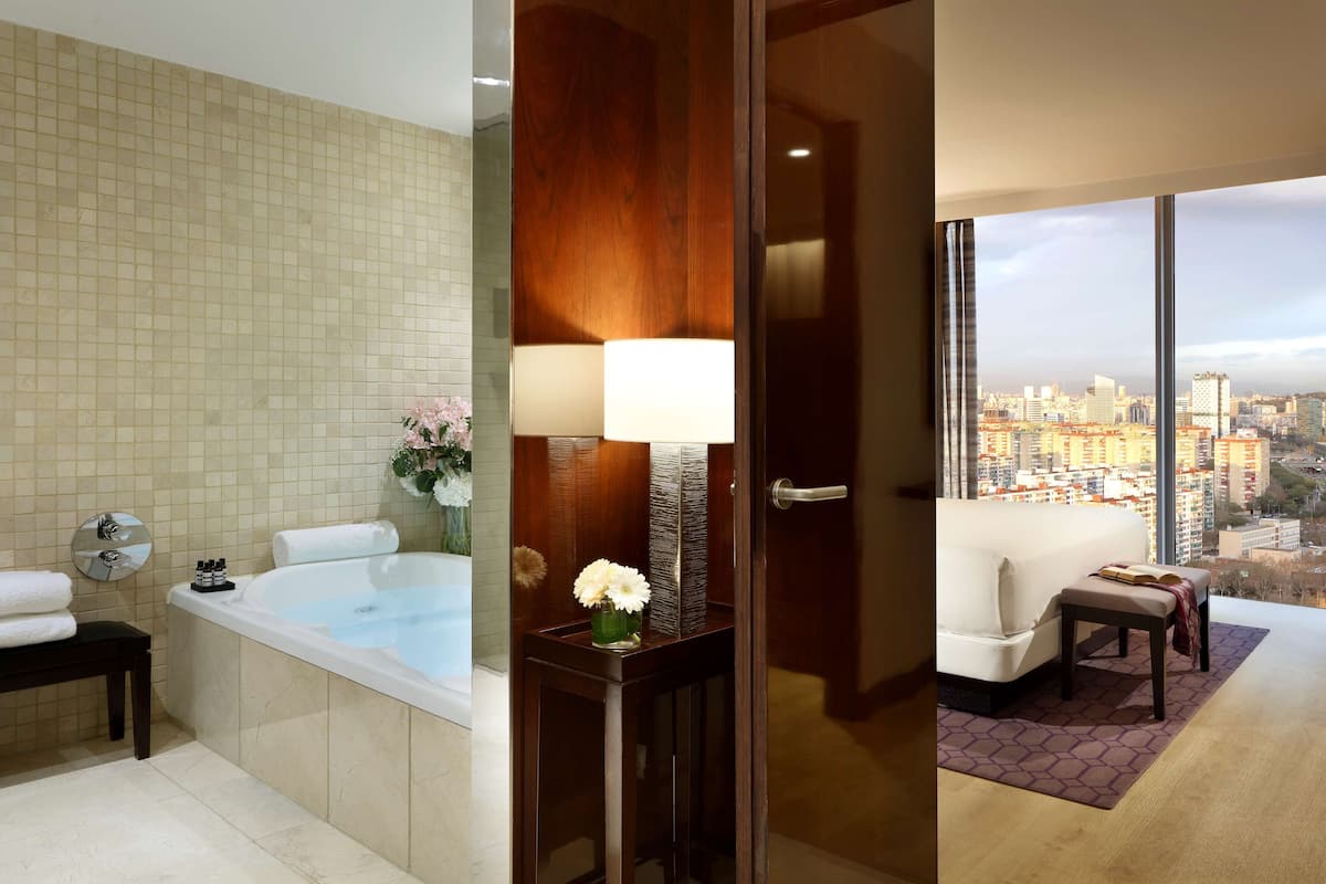 A hotel room with a bathtub, a bedside table with a lamp, a small table with a vase of flowers, and a view of the city through large windows.
