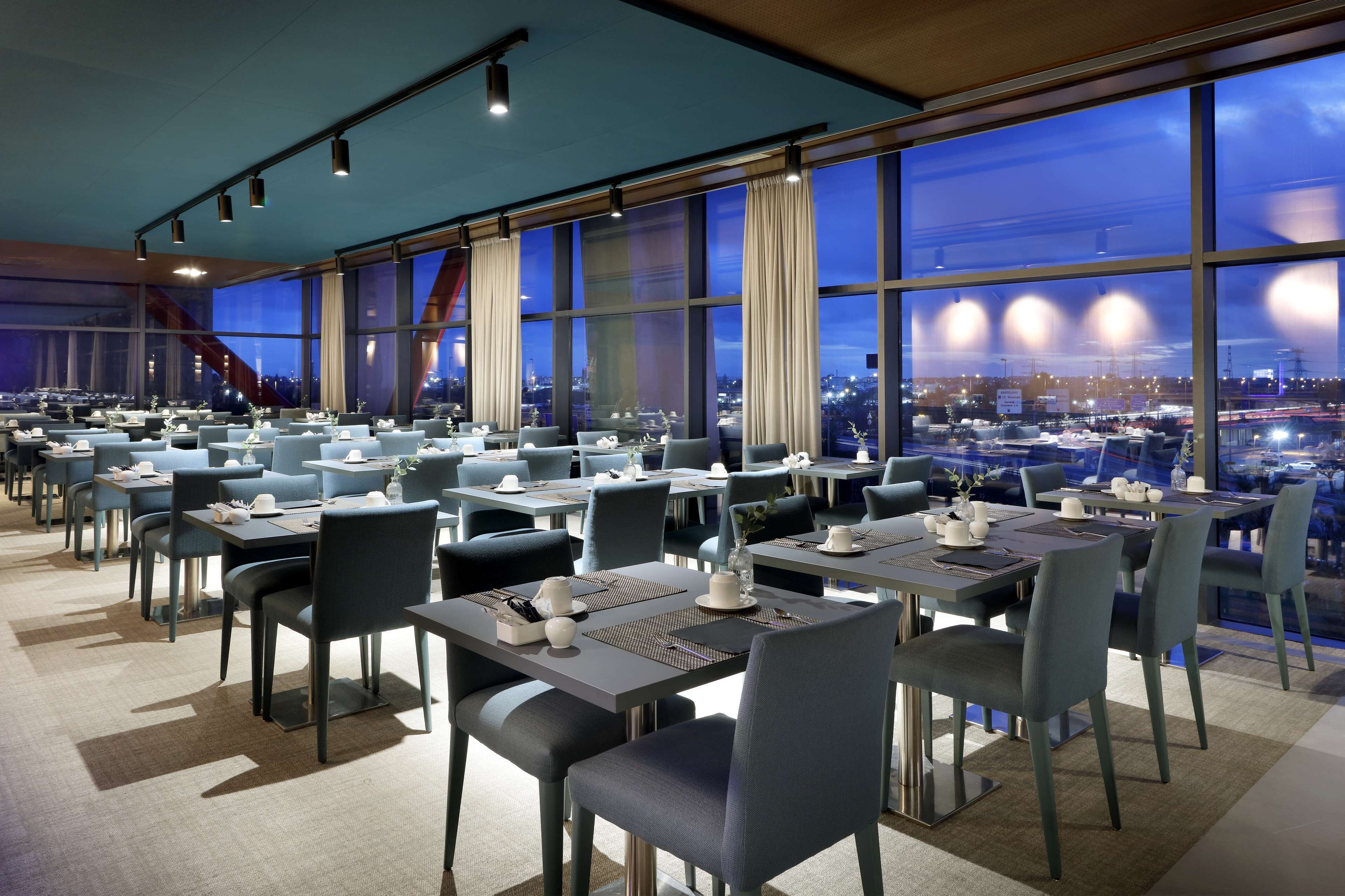 Un comedor moderno con mesas y sillas, ventanas amplias y vistas a la ciudad al anochecer.