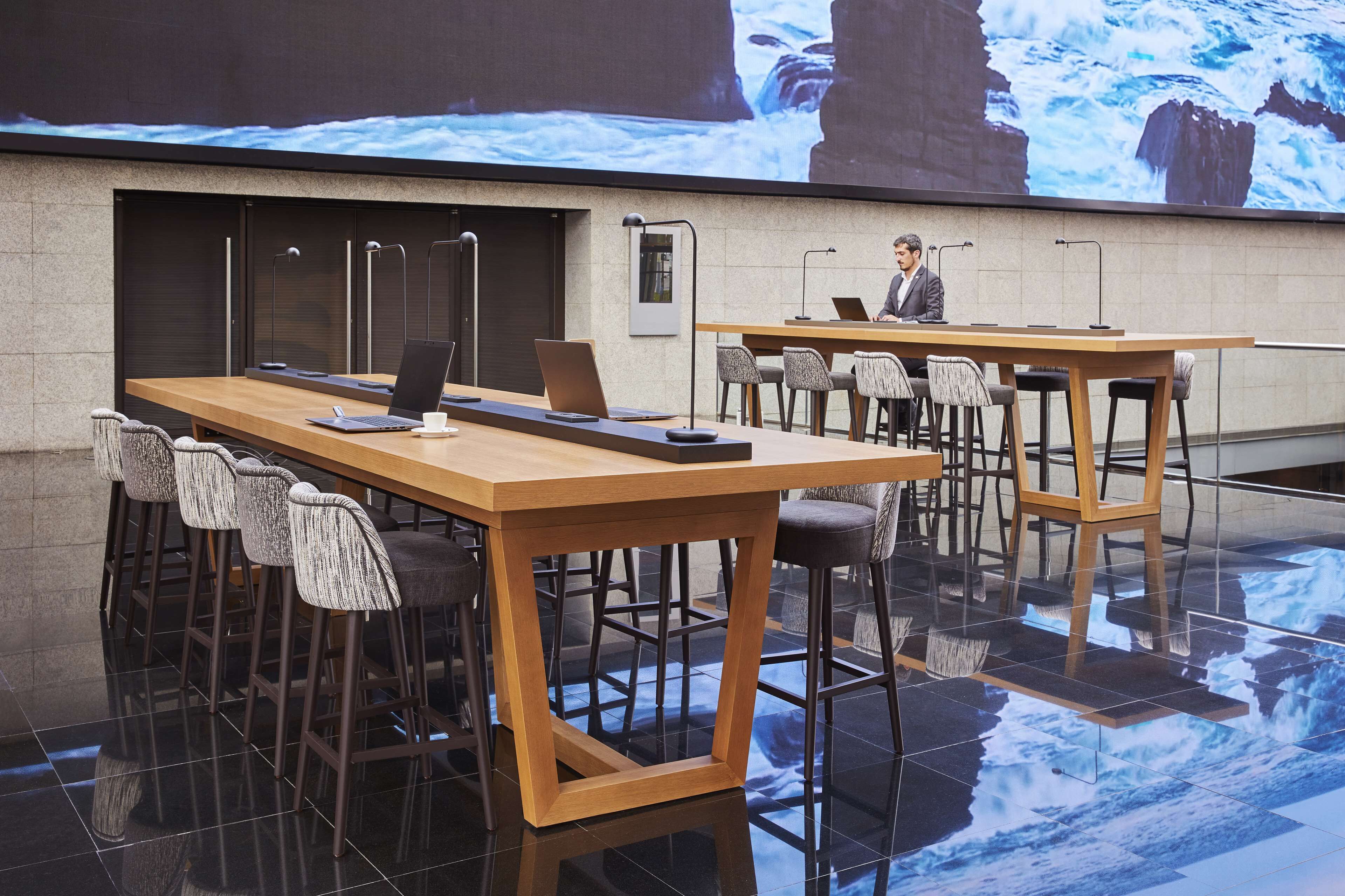 A modern conference room with a large screen displaying a scenic ocean view, wooden tables, and bar stools.