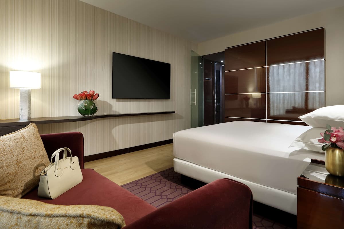 A hotel room with a bed, a TV mounted on the wall, a red sofa, a white handbag, and a vase with flowers.
