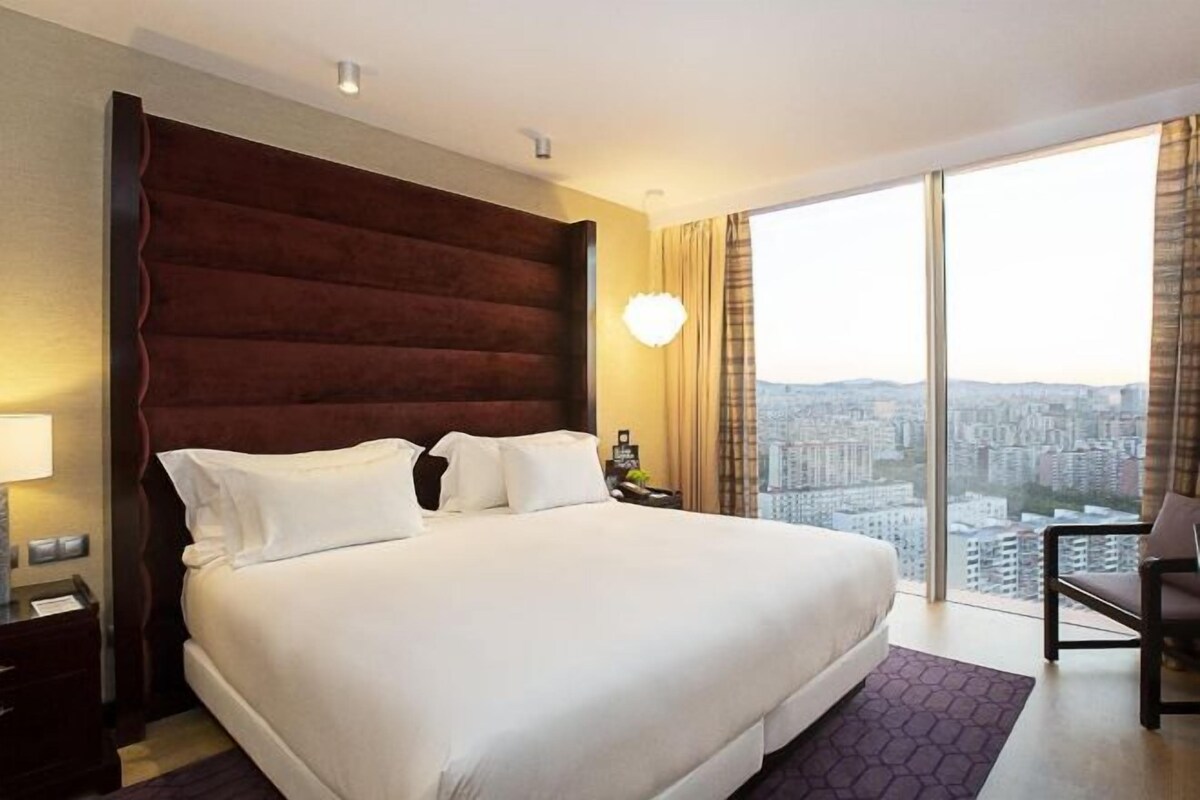 A hotel room with a large bed, a nightstand, a chair, and a view of the city through large windows.