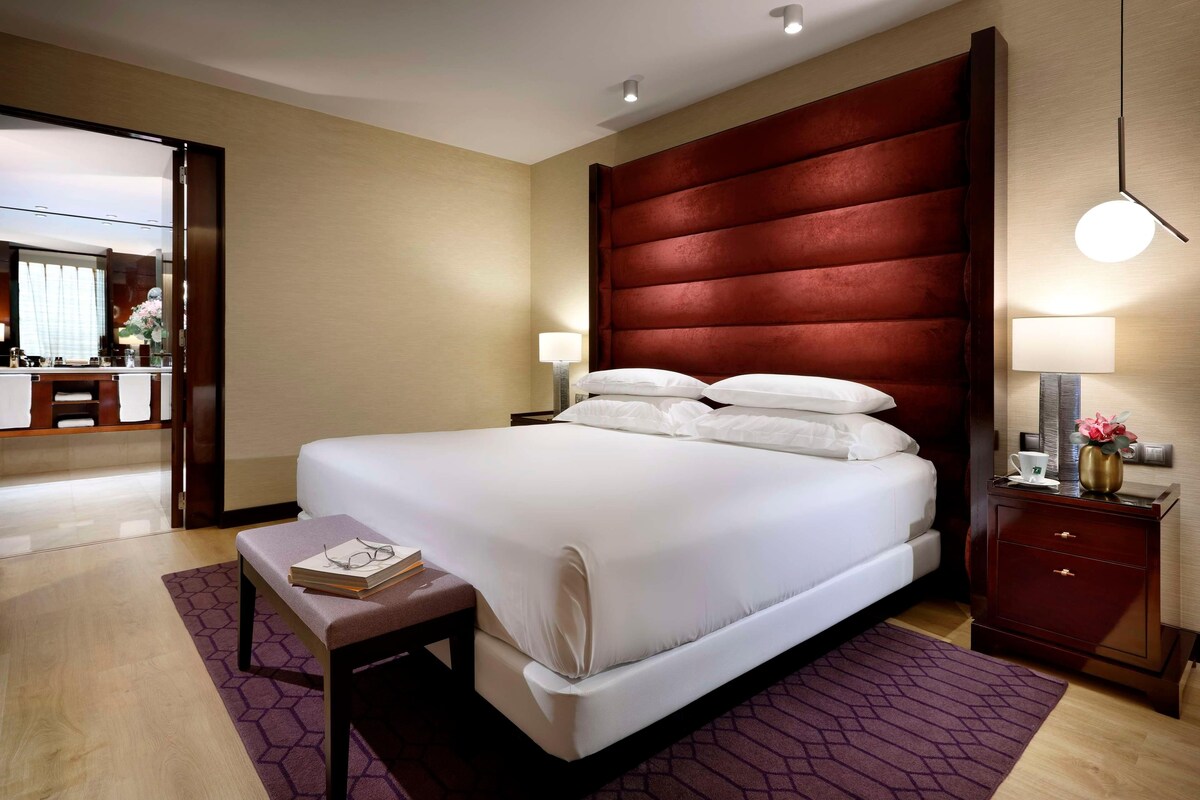 Ein Hotelzimmer mit einem großen Bett, einem Nachttisch mit Lampe, einem kleinen Nachttisch mit Vase sowie einem durchgegangenen Tür erkennbaren Badezimmer.
