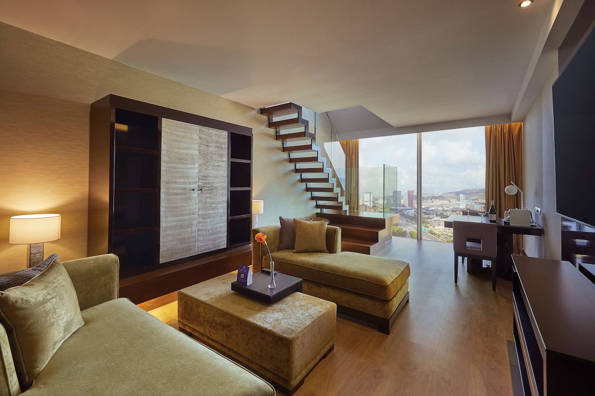 A modern hotel room with a staircase, a large window with a city view, a sofa, a coffee table, and a desk with a lamp.