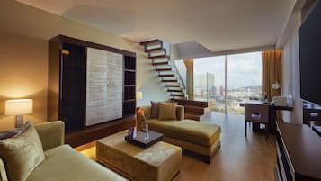 Una habitación de hotel moderna con una escalera, un ventanal con vista a la ciudad, un sofá, una mesa de centro y un escritorio con una lámpara.