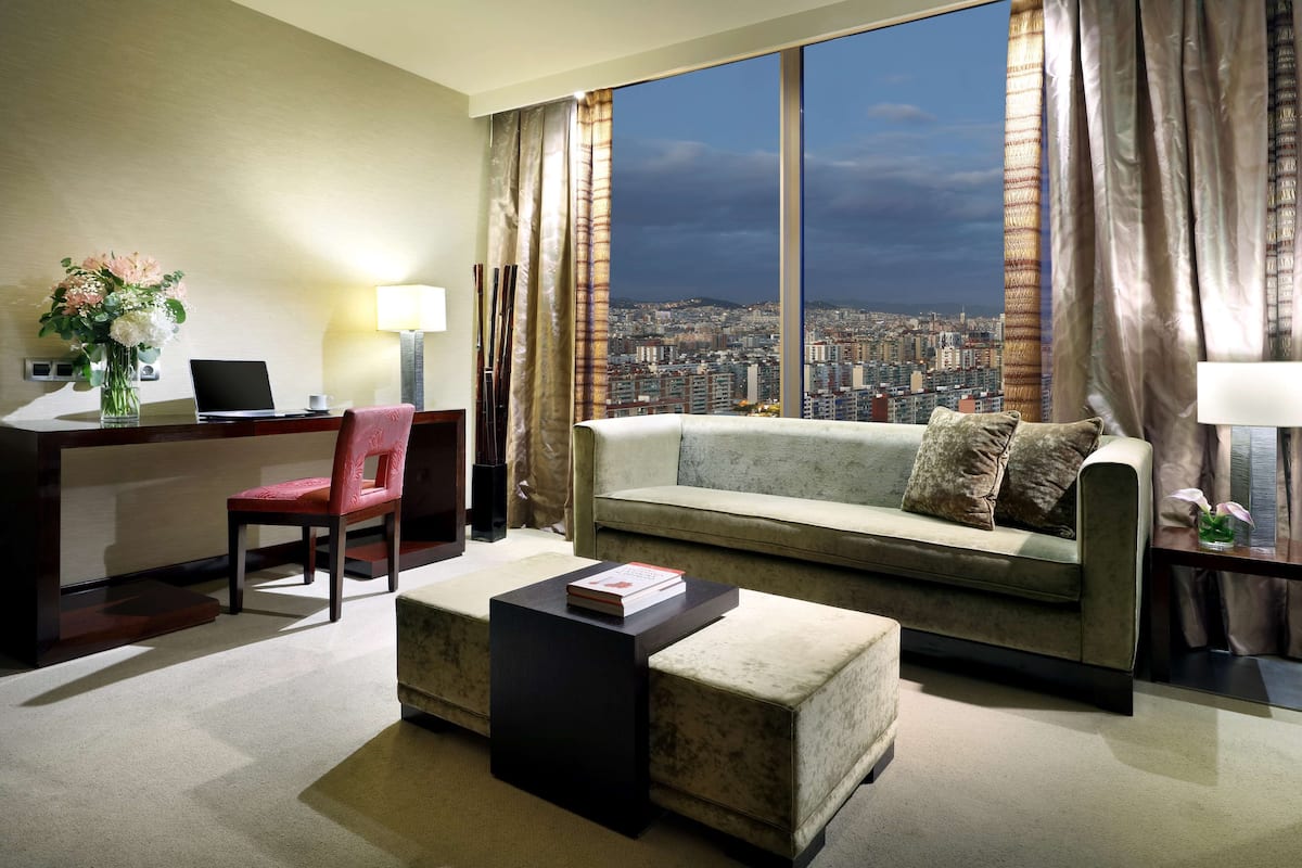 A modern living room with a city view, a desk with a laptop, a red chair, a grey sofa, and a small black ottoman.