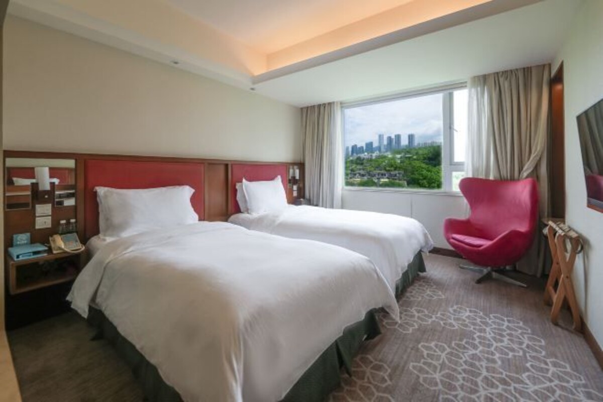 Une chambre d’hôtel avec deux lits, une chaise rouge, une fenêtre donnant sur la ville et une table de chevet en bois avec un téléphone et une lampe.