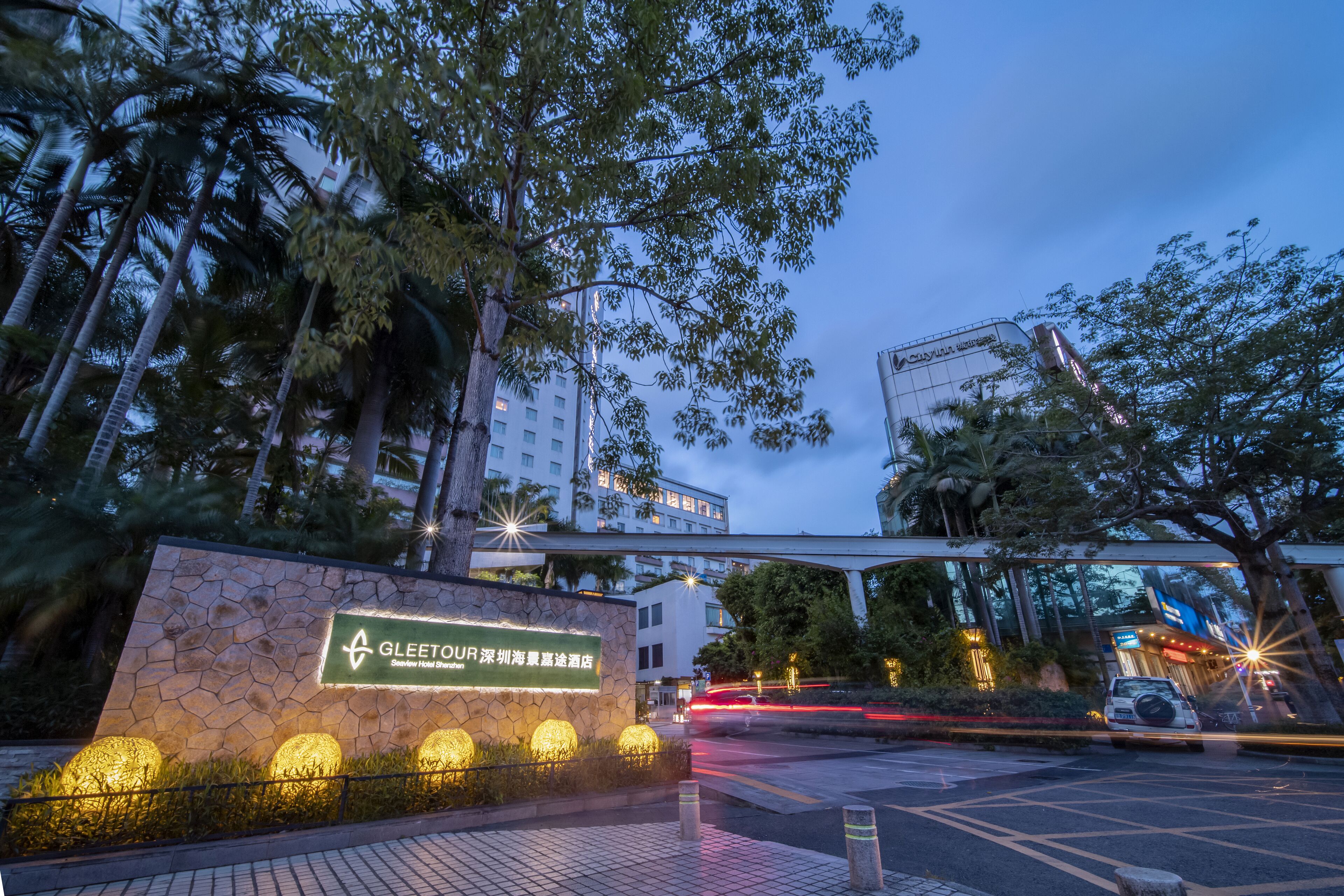 A hotel entrance with a stone facade, illuminated sign, and a pedestrian overpass above.