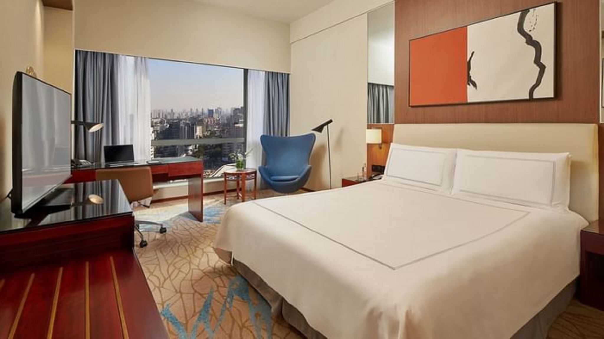 A hotel room with a large bed, a desk with a computer, a chair, and a view of the city.