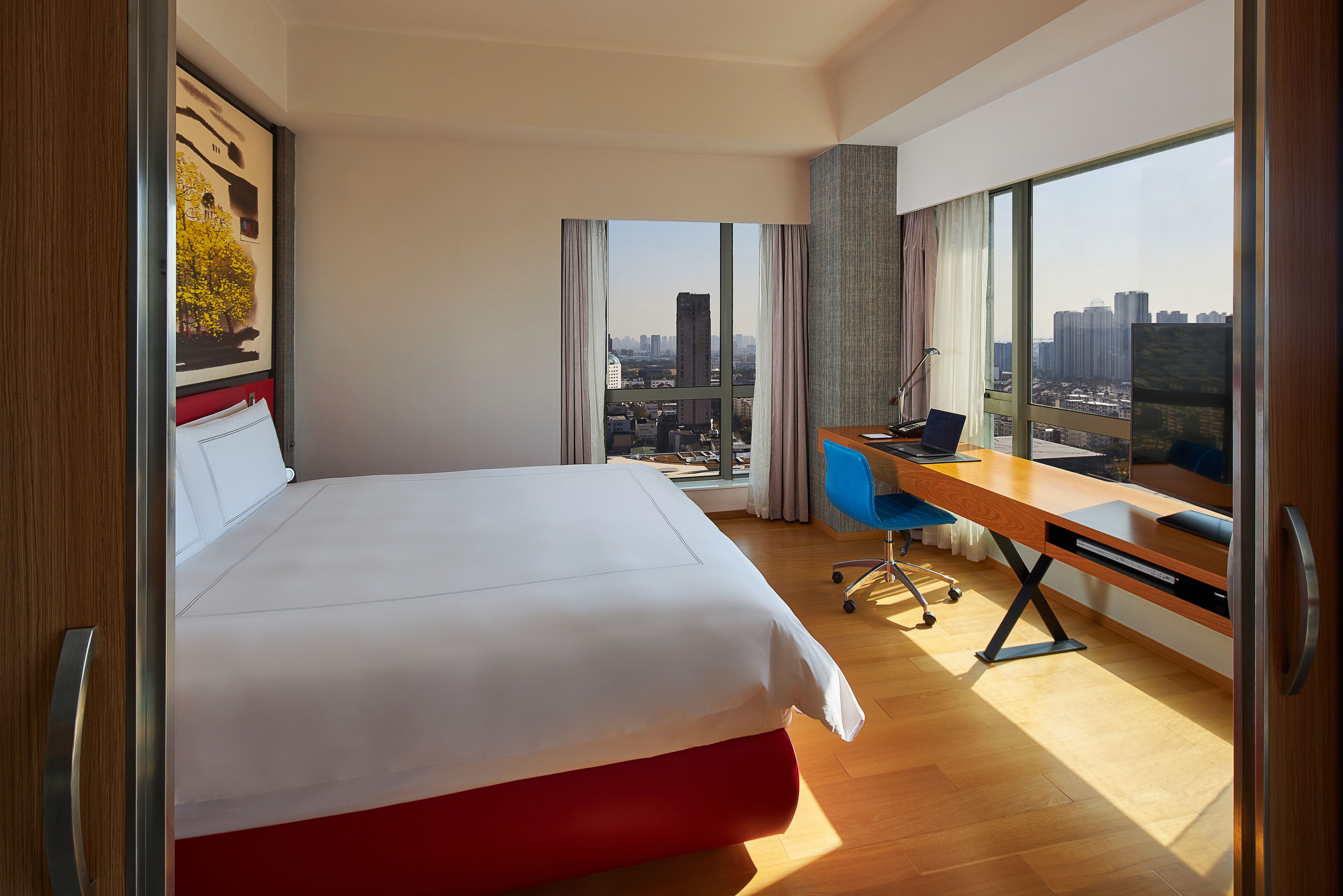 A hotel room with a large bed, a desk with a laptop, and a view of a cityscape through large windows.