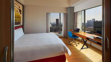 A hotel room with a large bed, a desk with a laptop, and a view of a cityscape through large windows.