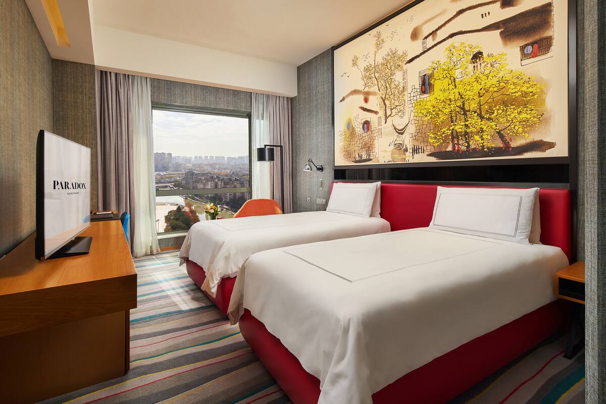 A hotel room with two beds, a desk, and a large window offering a city view.
