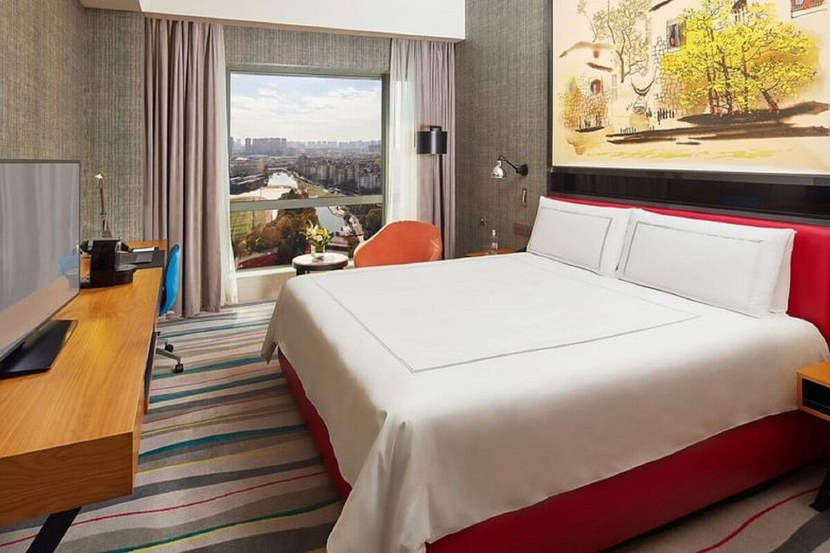 A hotel room with a large bed, a desk with a computer, a chair, a small table with a vase, and a window with a city view.