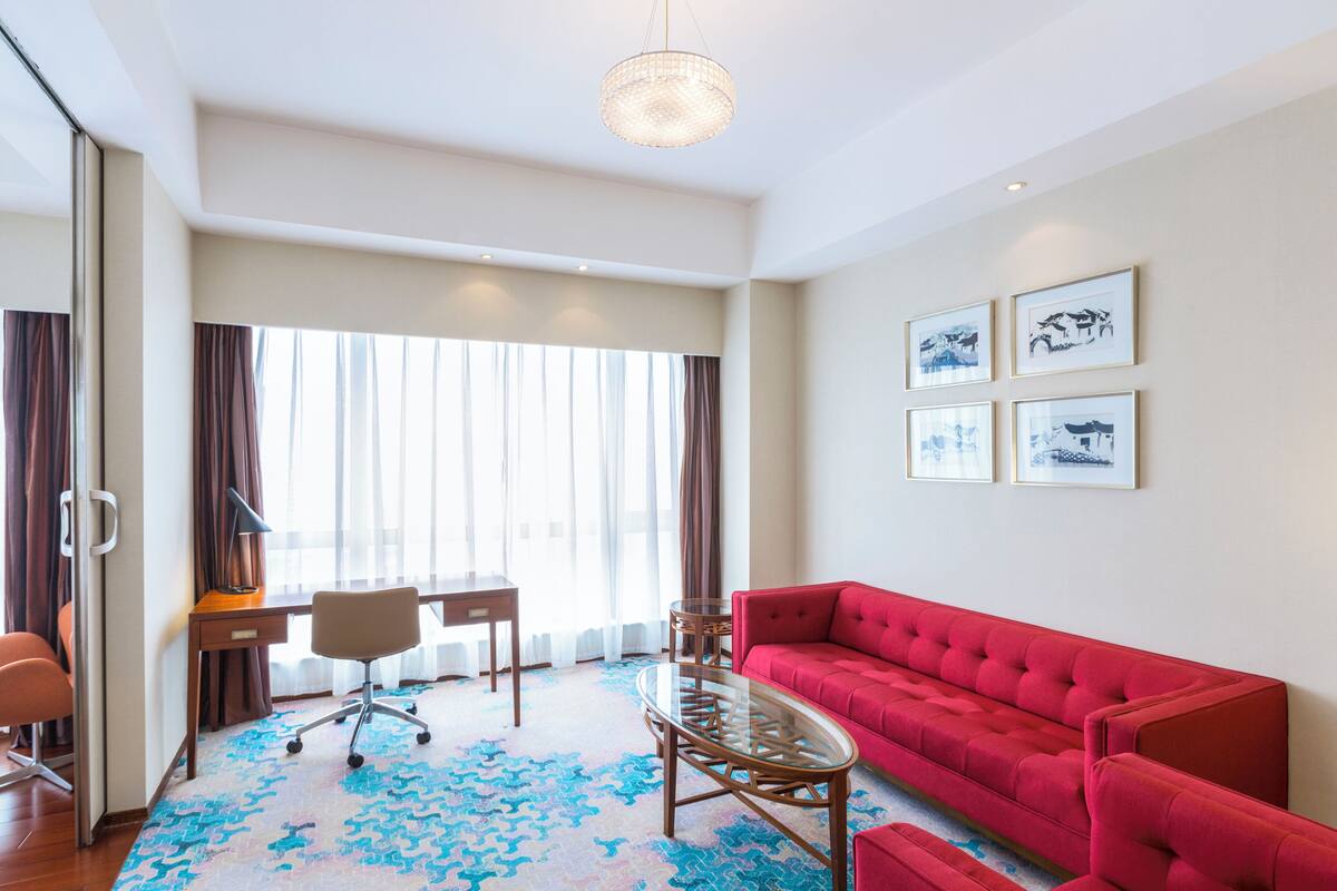 A modern hotel room with a red sofa, a glass coffee table, a desk, and a chair. There are large windows with curtains, and the room has a light-colored carpet.