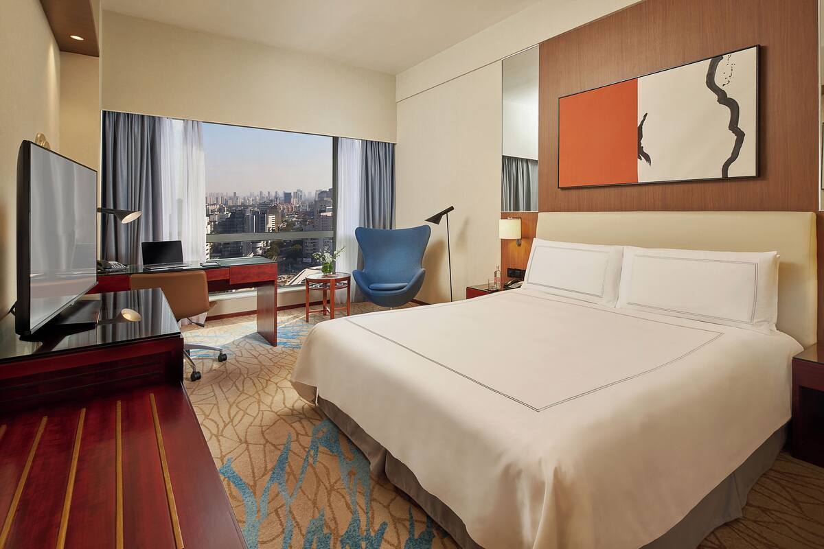 A hotel room with a large bed, a desk with a laptop, a chair, and a view of the city.