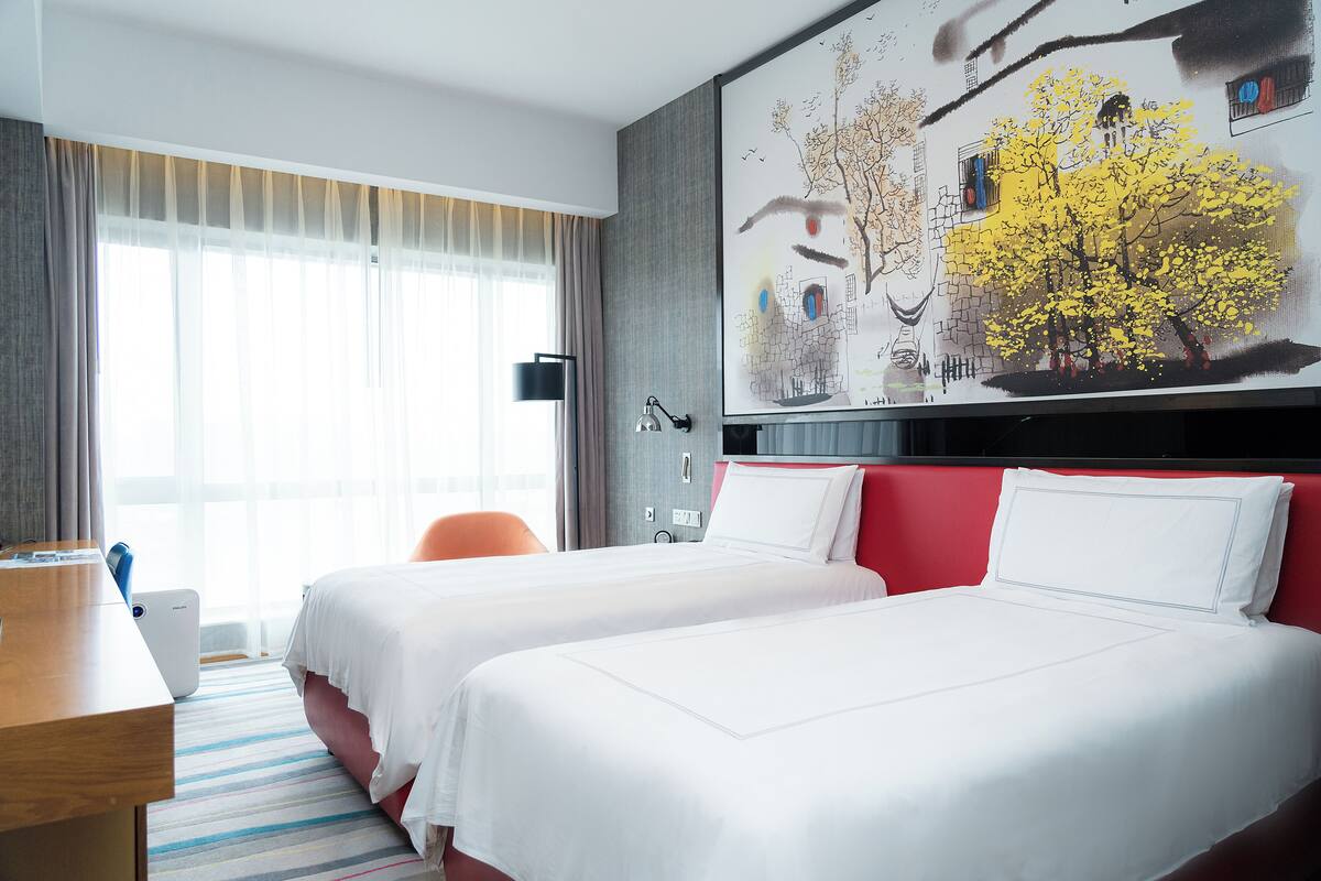 A hotel room with two beds, a large window with sheer curtains, a painting on the wall, and a desk with a lamp.