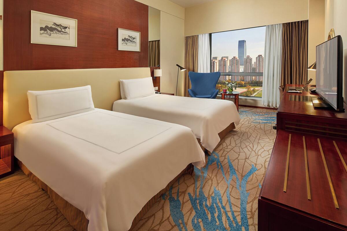 A hotel room with two beds, a desk with a TV, a chair, and a large window with a city view.