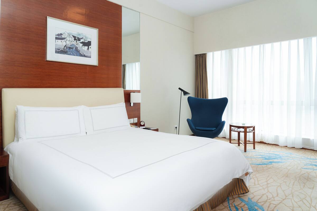 A hotel room with a large bed, a blue armchair, a small table, and a painting on the wall.