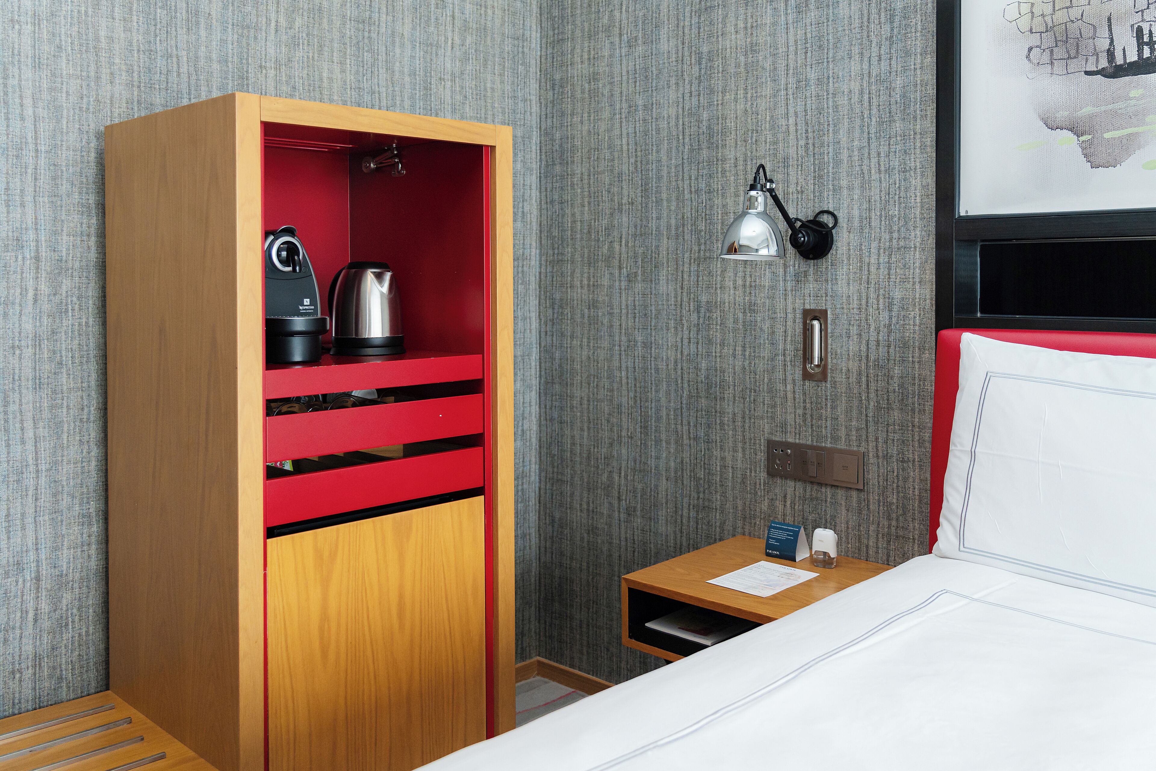 A hotel room with a wooden wardrobe, a red interior, a bed with white bedding, a nightstand, a lamp, and a map on the wall.