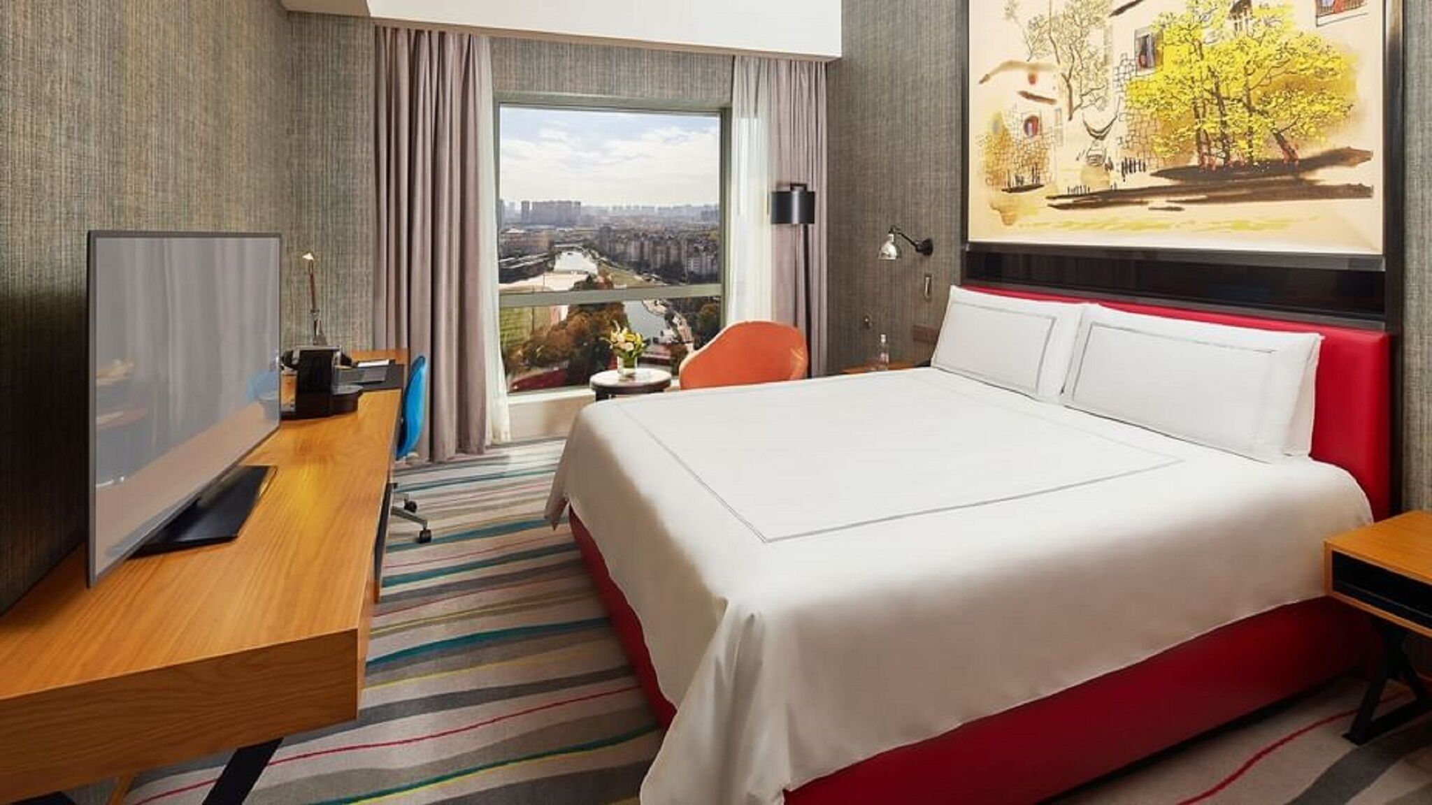 A hotel room with a large bed, a desk with a computer, a chair, a small table with a vase, and a window with a city view.