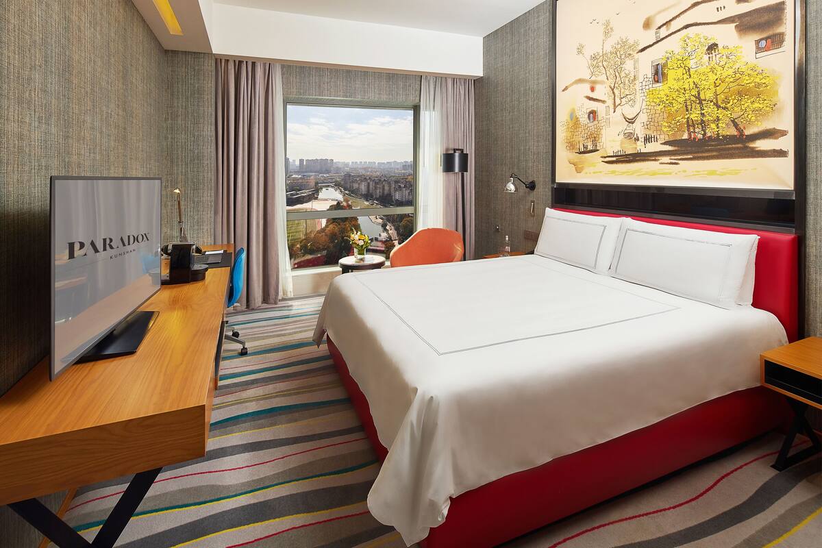 A hotel room with a large bed, a desk with a computer, a TV, and a view of a cityscape.