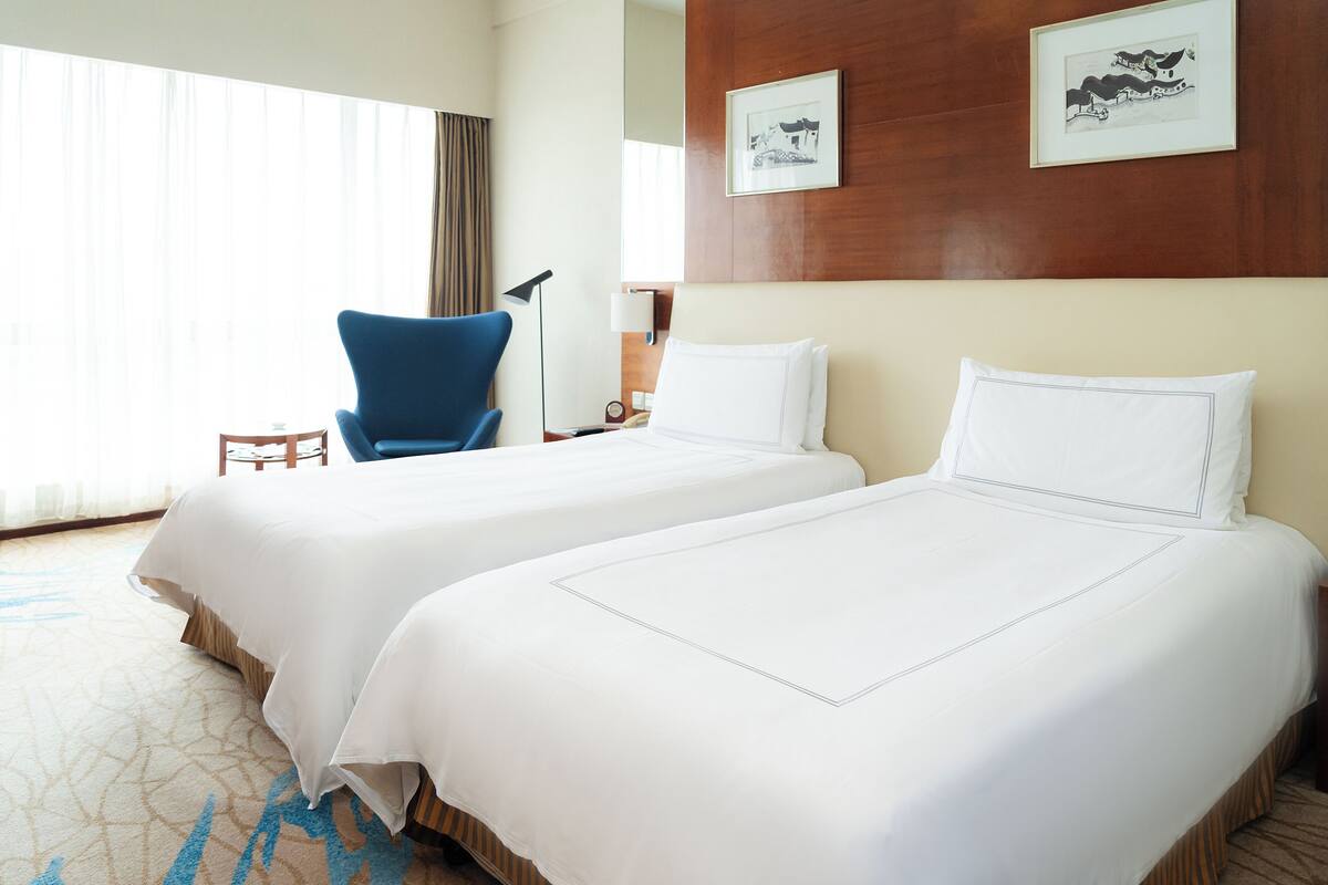 A hotel room with two beds, a blue chair, a small table, and a lamp.