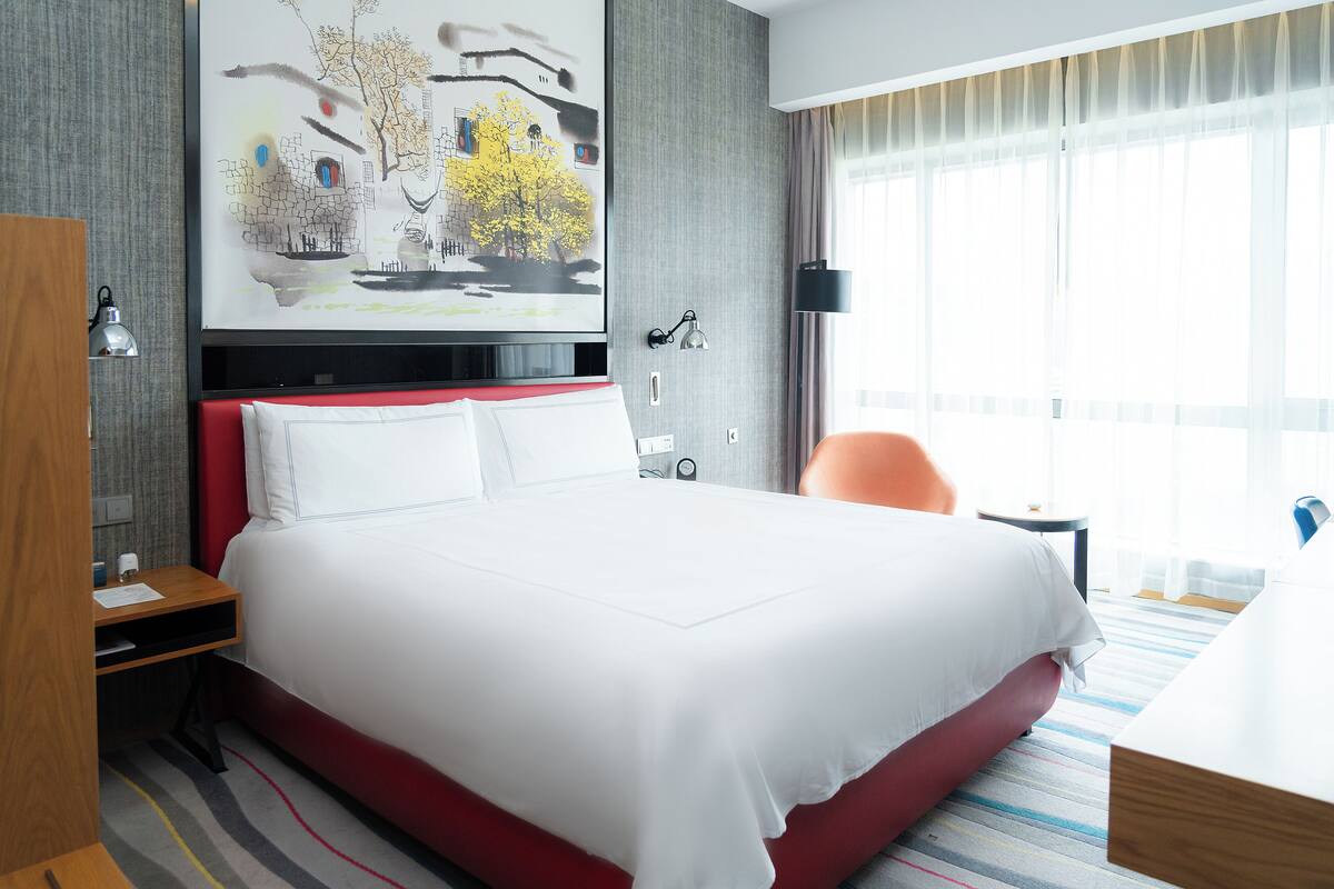 A hotel room with a large bed, a desk, a chair, a painting on the wall, and a window with curtains.
