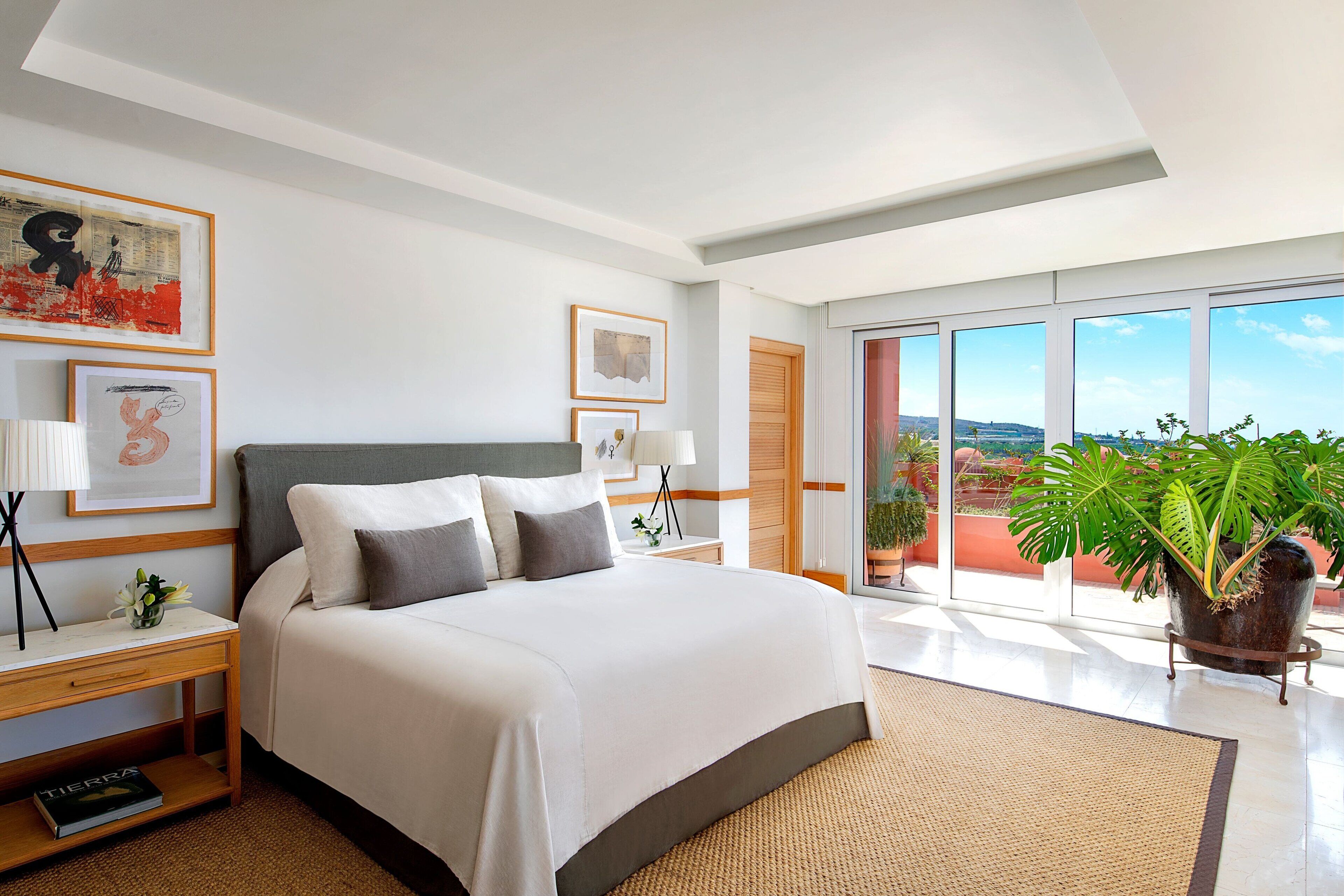 Une chambre d’hôtel moderne avec un grand lit, une table de chevet, un bureau, une plante en pot et une vue sur la ville grâce à de grandes fenêtres.