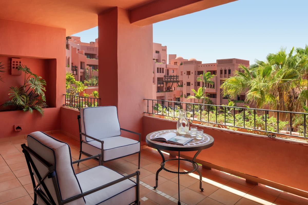 Un balcón con dos sillas y una mesita, con vistas a una zona residencial con palmeras y edificios.