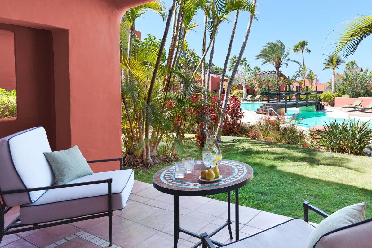 Un patio con una silla lounge blanca, una pequeña mesa redonda con un florero y fruta, y vistas a una piscina y palmeras.