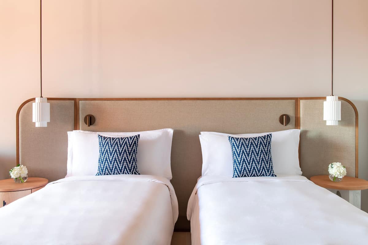 Habitación de hotel con dos camas, cada una con ropa de cama blanca y almohadas azules estampadas. Hay dos mesitas de noche con lámparas blancas y pequeños arreglos florales.
