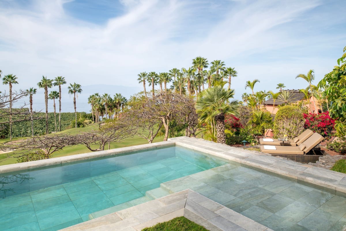Piscina con agua cristalina, rodeada de palmeras y un área de descanso con una silla y una mesa.