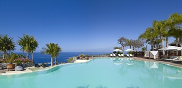 A pool with lounge chairs and umbrellas, palm trees, and a view of the ocean.