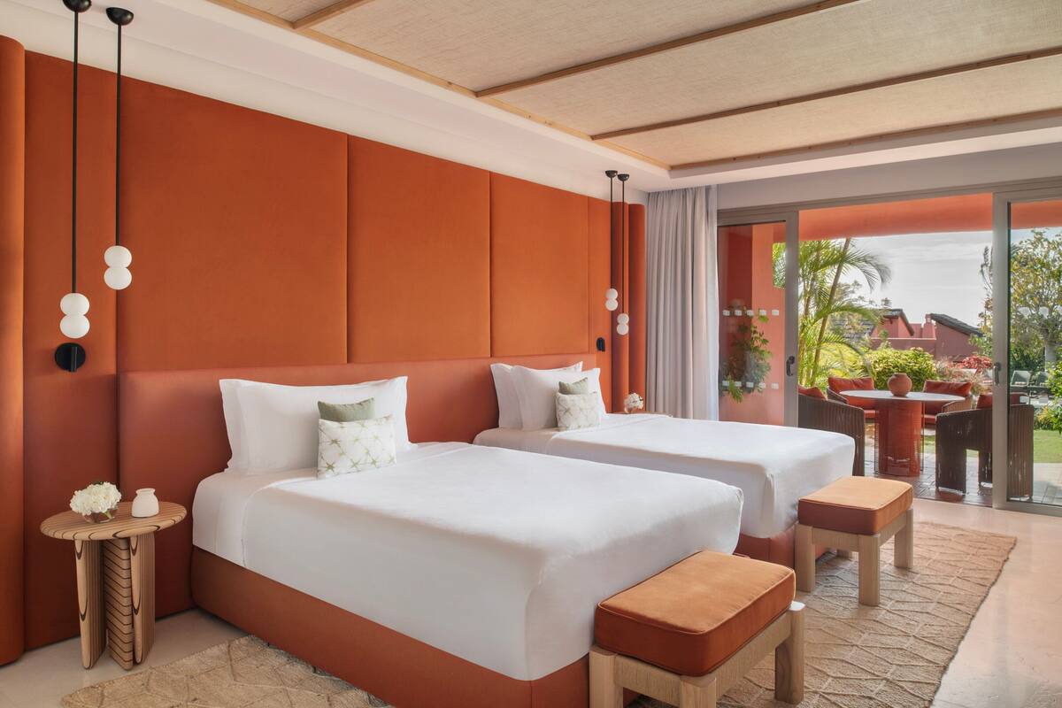 Habitación de hotel con dos camas, una mesita con un florero y vistas al exterior.