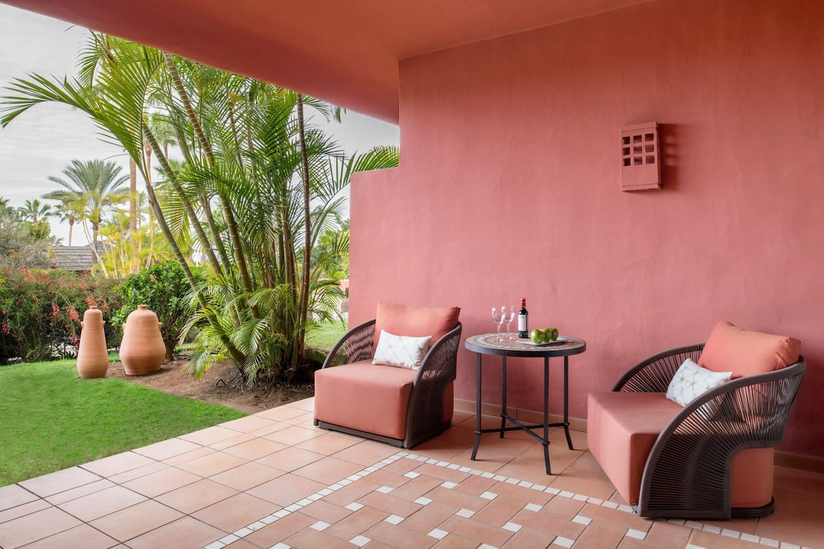 Un patio con dos sillas de mimbre, una mesita redonda y una botella de vino. Se observan macetas de terracota grandes y palmeras al fondo.